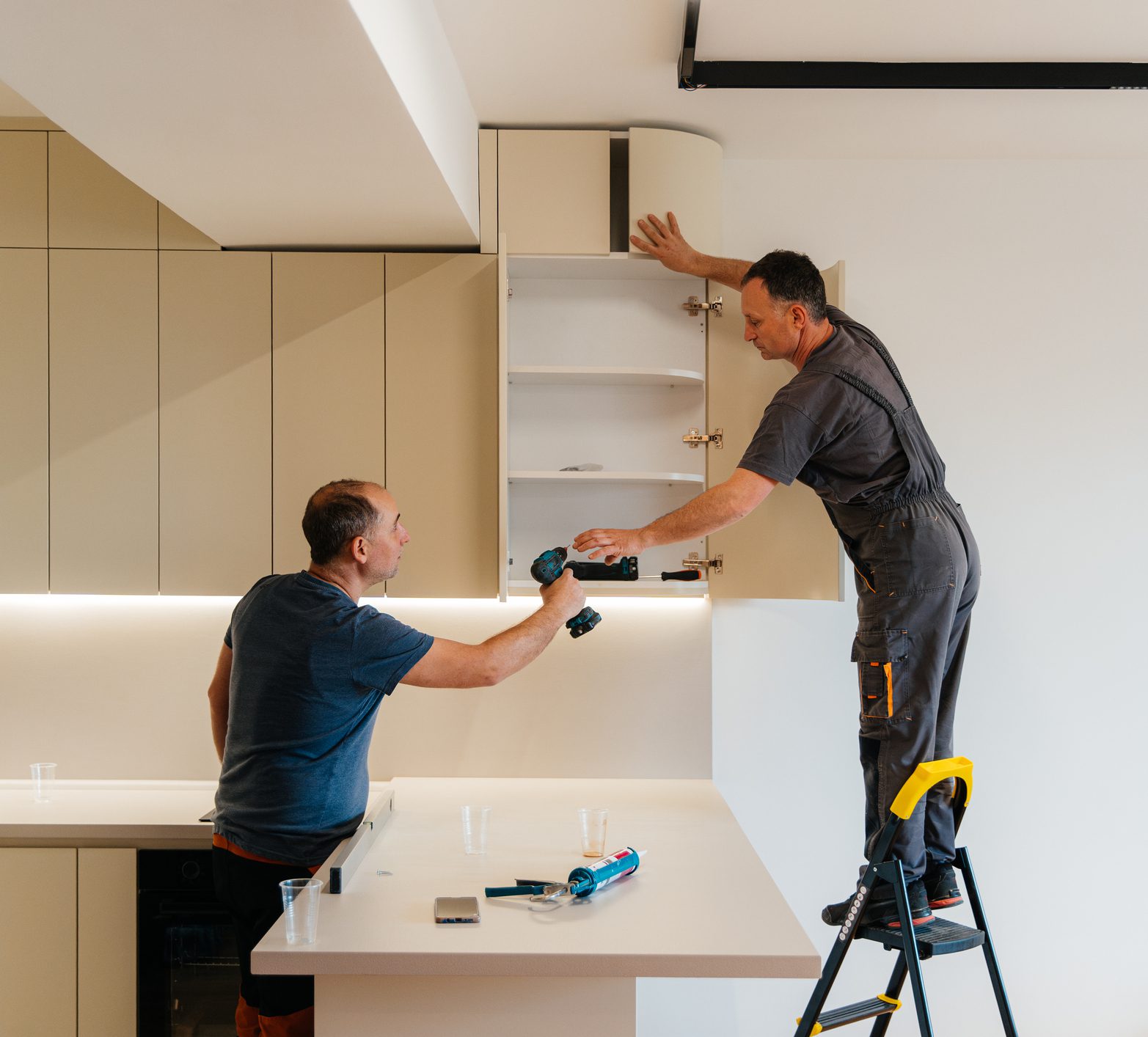 Two male workers collaborate while installing upper kitchen cabinets in a modern kitchen. One man stands on a step ladder holding the cabinet door, while the other hands him a cordless drill across the kitchen counter. The bright interior, clean surfaces, and tools on the countertop highlight a professional kitchen installation process.